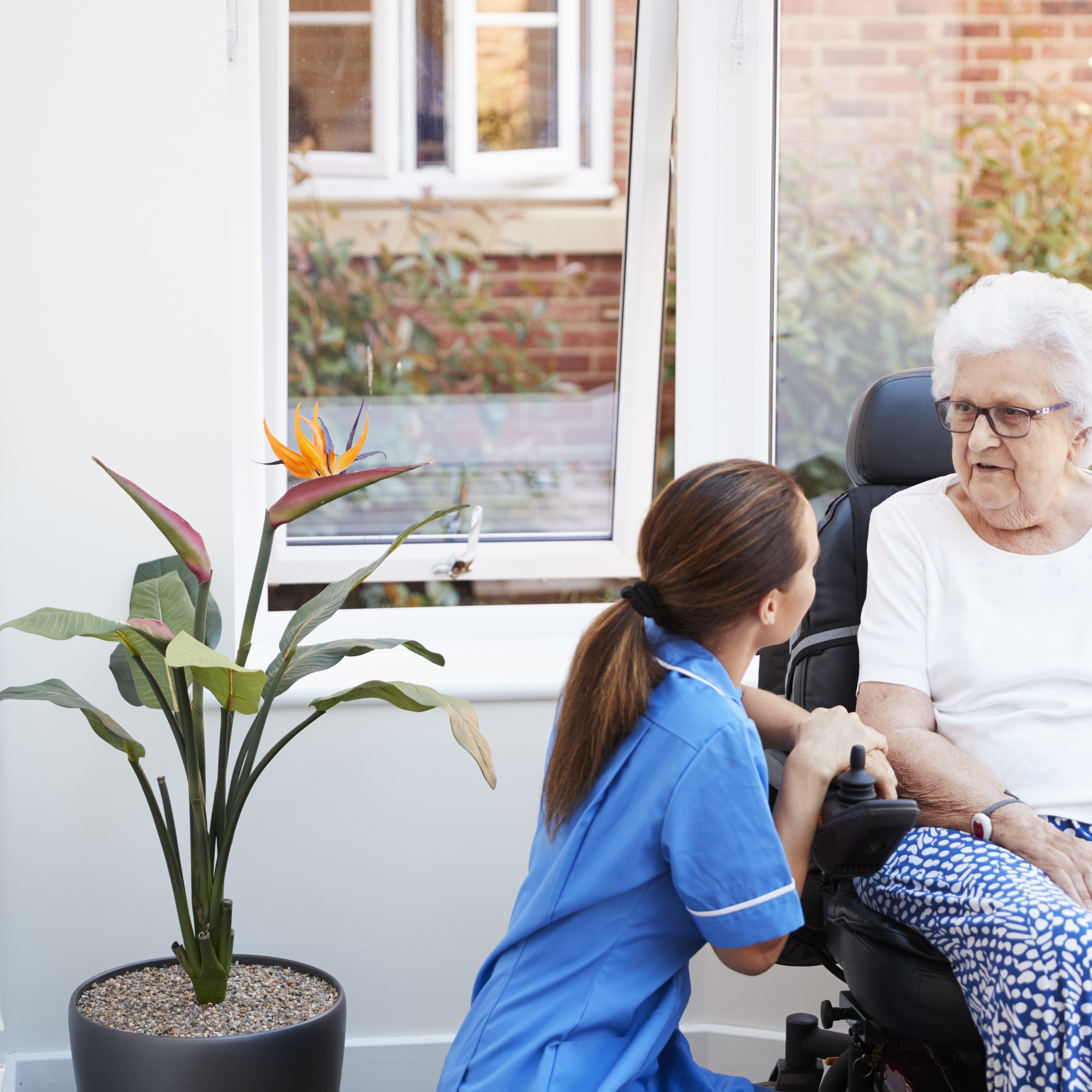 Senior Woman Sitting In Motorized Wheelchair Talking With Nurse In Retirement Home